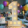 A large illuminated number one marquee sign made of LED lights stands on a table surrounded by balloons and birthday decorati