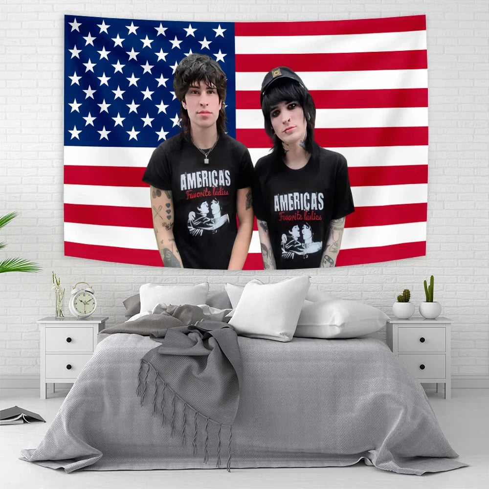 A portrait of two people standing in front of an American flag tapestry hanging on a white brick wall.