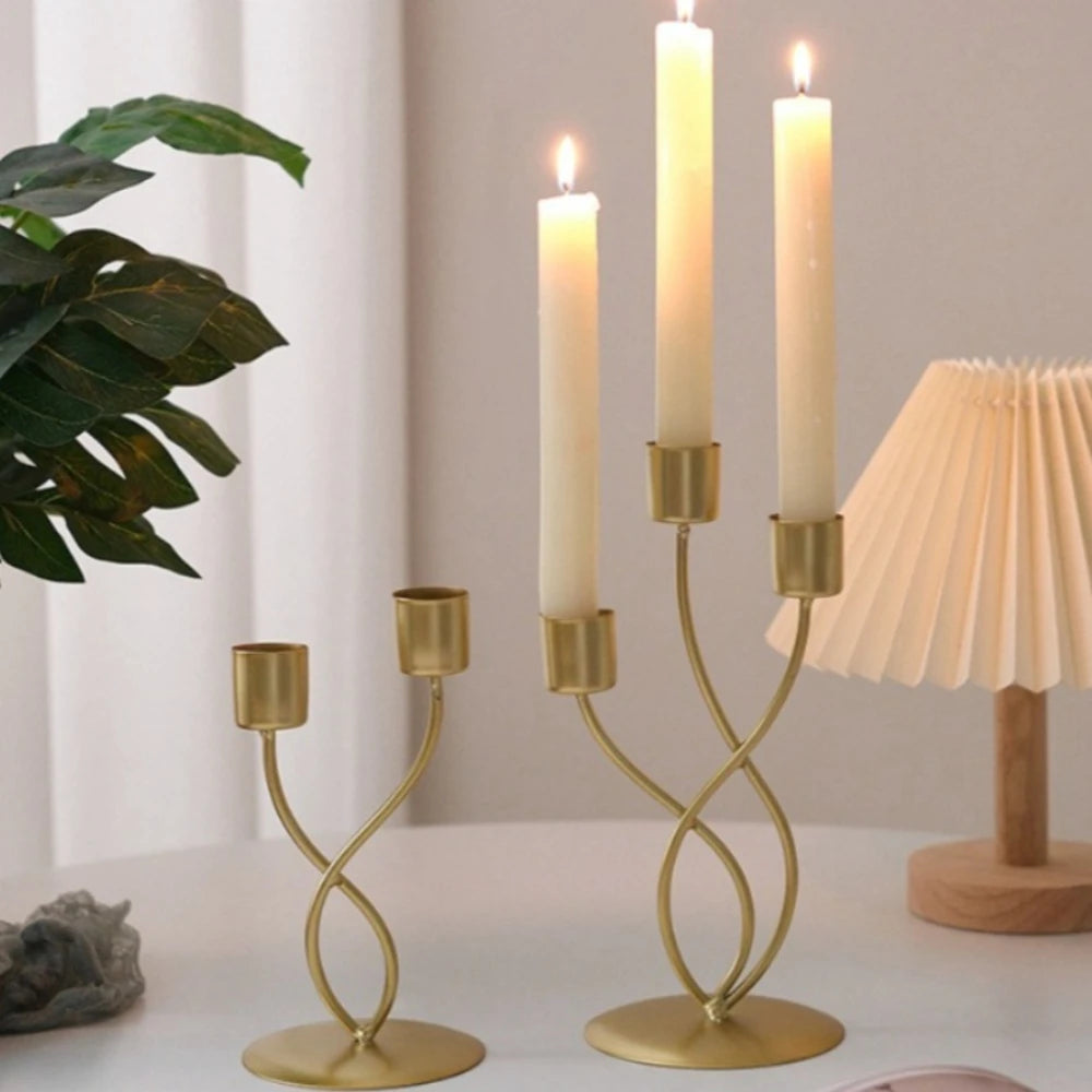 A gold iron candle holder with slender white candles on a white table next to a green plant and a lamp.