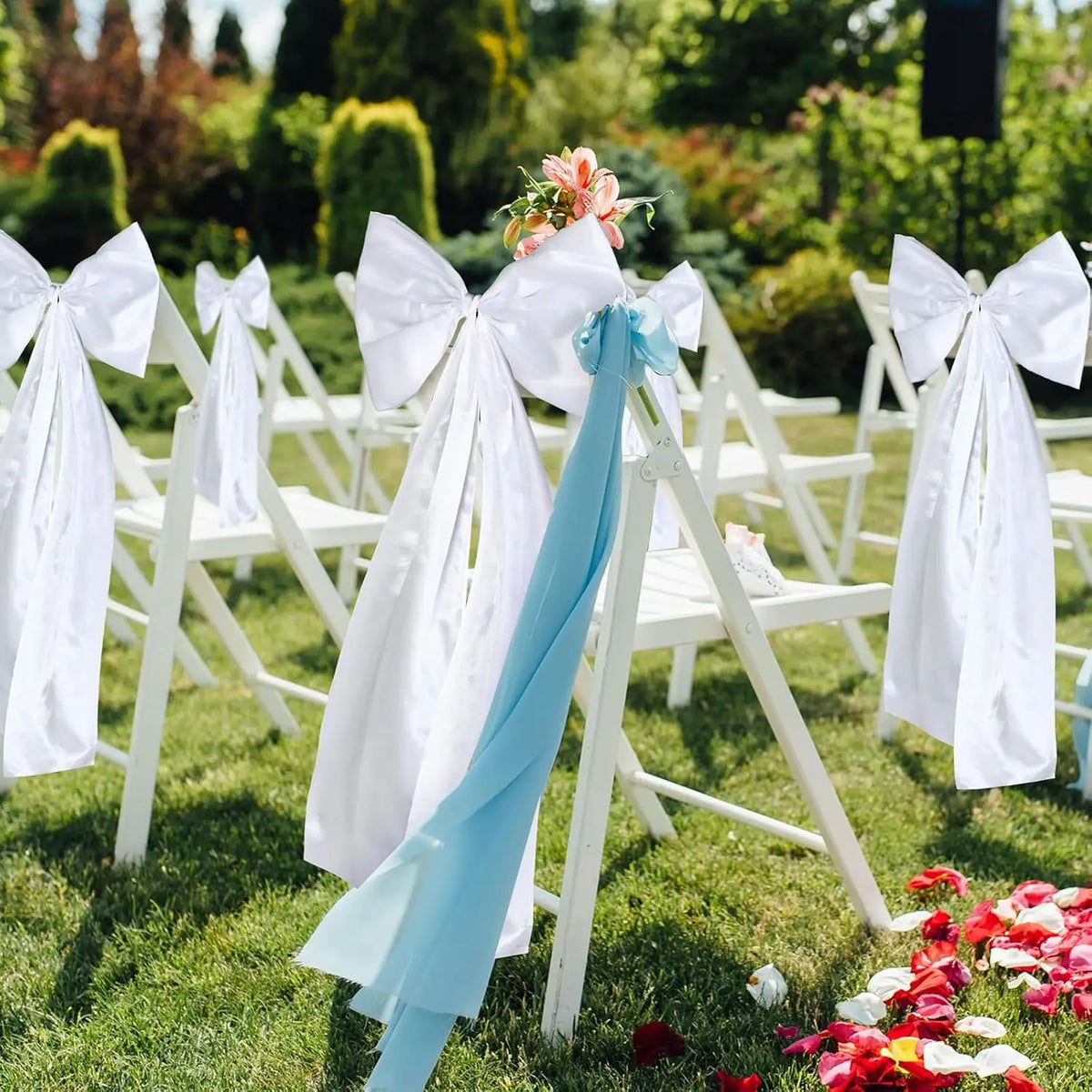 Nœuds de satin blanc et bleu clair pour mariage attachés sur des chaises blanches dans un jardin vert.