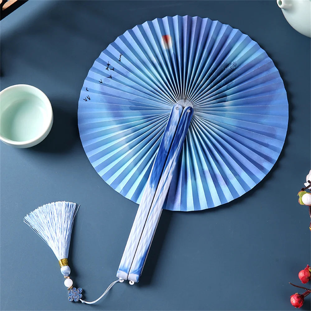 A blue and white traditional Chinese folding fan with a tassel resting on a dark blue surface.