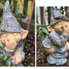 A gray gnome statue with a pointed hat and rosy cheeks is nestled among green foliage.