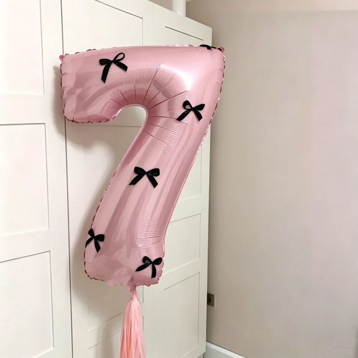 A pink foil balloon shaped like the number 7 with black bows, hanging on a white door.
