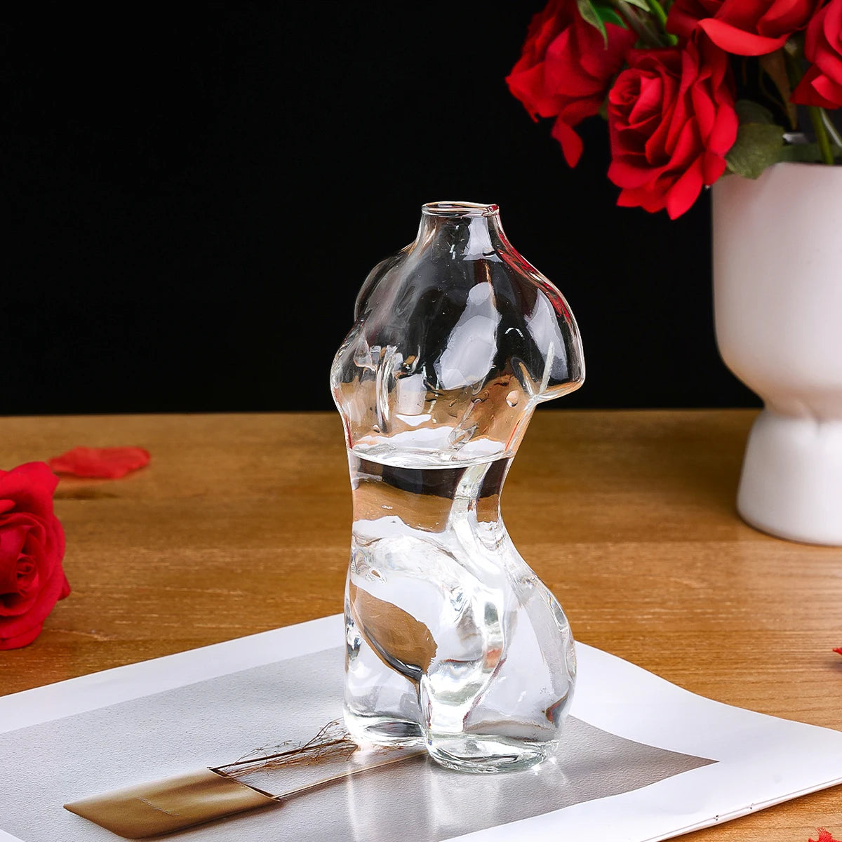 Artistic glass human body vase filled with water on a wooden surface next to red roses