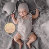 A baby wearing a knitted outfit is lying on a gray blanket with a wooden name announcement sign nearby.