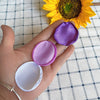 A hand holds several shades of purple and white silk rose petals on a checkered cloth next to a sunflower.