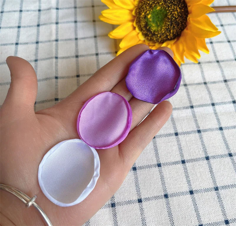 A hand holds several shades of purple and white silk rose petals on a checkered cloth next to a sunflower.