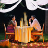 A couple is seated at a table decorated with tall acrylic candelabra outdoors surrounded by pink flowers.