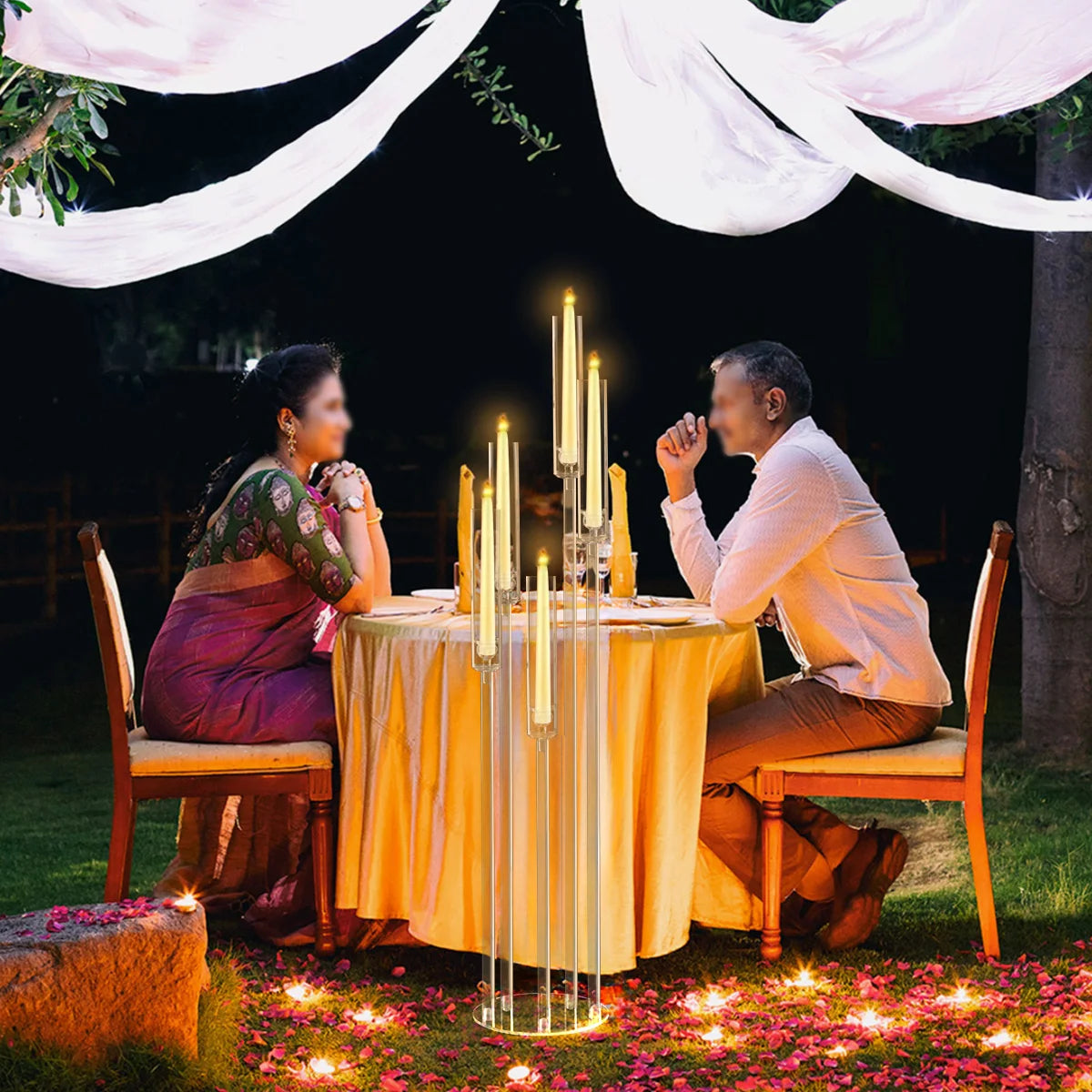 A couple is seated at a table decorated with tall acrylic candelabra outdoors surrounded by pink flowers.