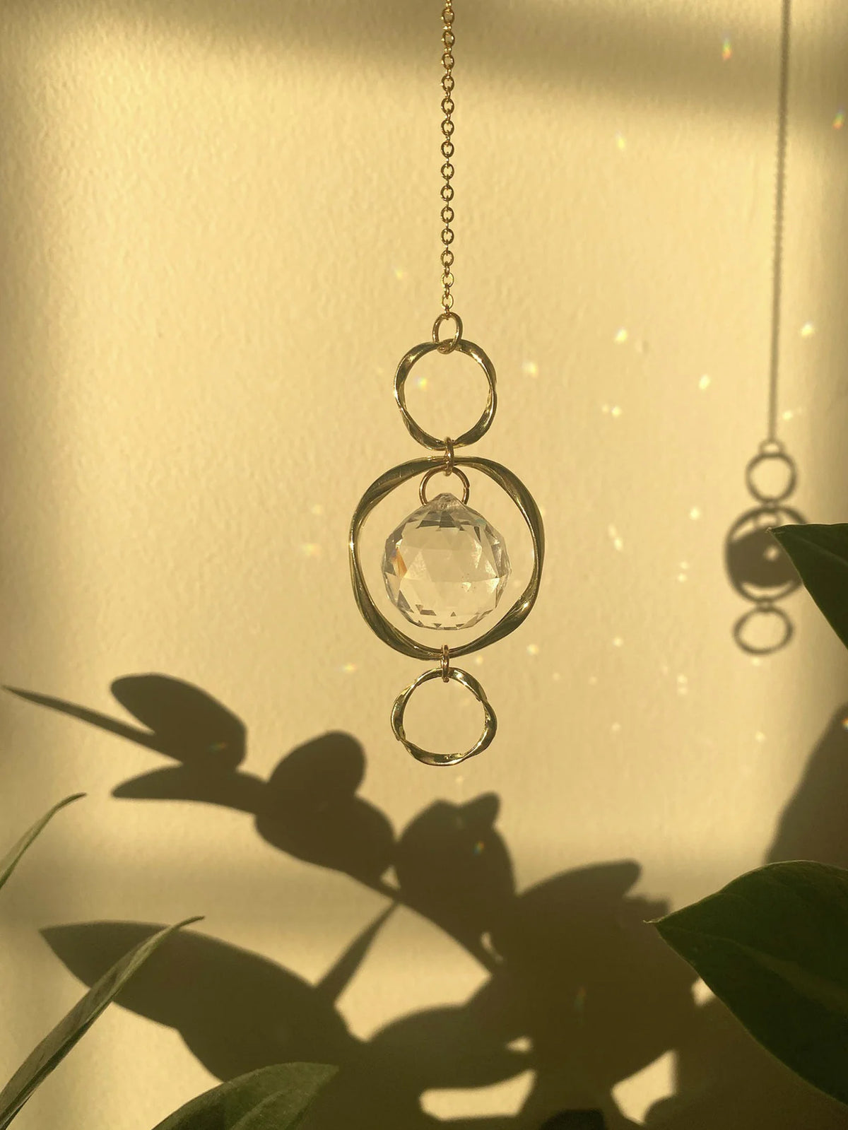 Une délicate chaîne en or avec un pendentif de cristal clair à anneaux concentriques contre un mur ensoleillé.