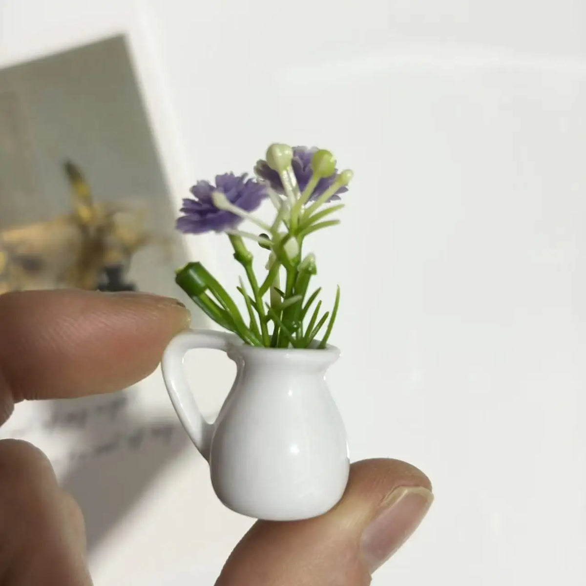 A white ceramic vase holding artificial purple flowers, being held by a hand.