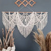 Handwoven boho macrame wall hanging tapestry with white fringe suspended on a wooden rod against a gray wall.