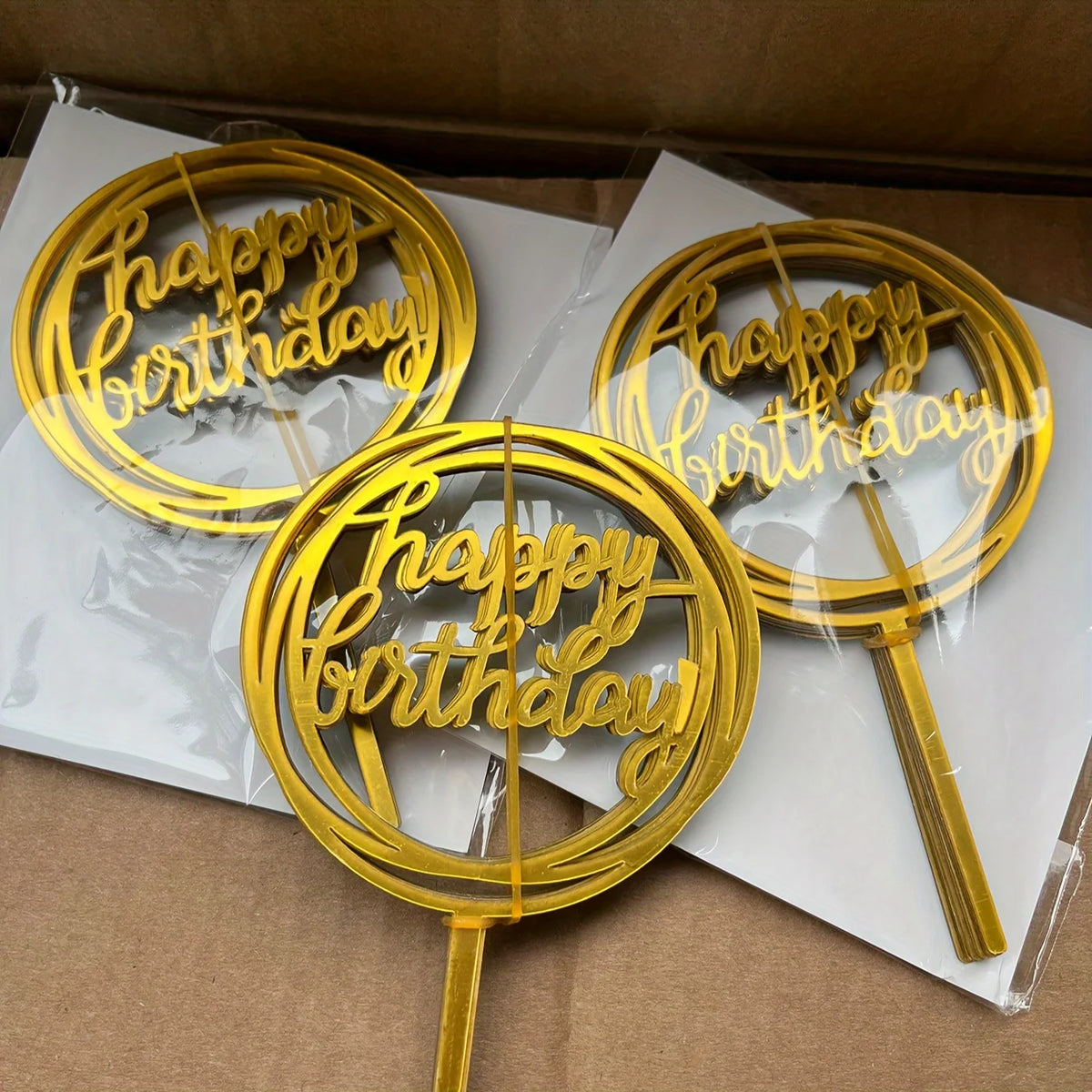 A set of ten gold acrylic happy birthday cake toppers are displayed in a cardboard box.