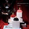 A display featuring a beautiful low temperature rose candle surrounded by dark floral elements and stacked white boxes.