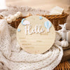 A round wooden birth announcement sign with the word Hello and a cloud and hot air balloon design, resting on a white knitted