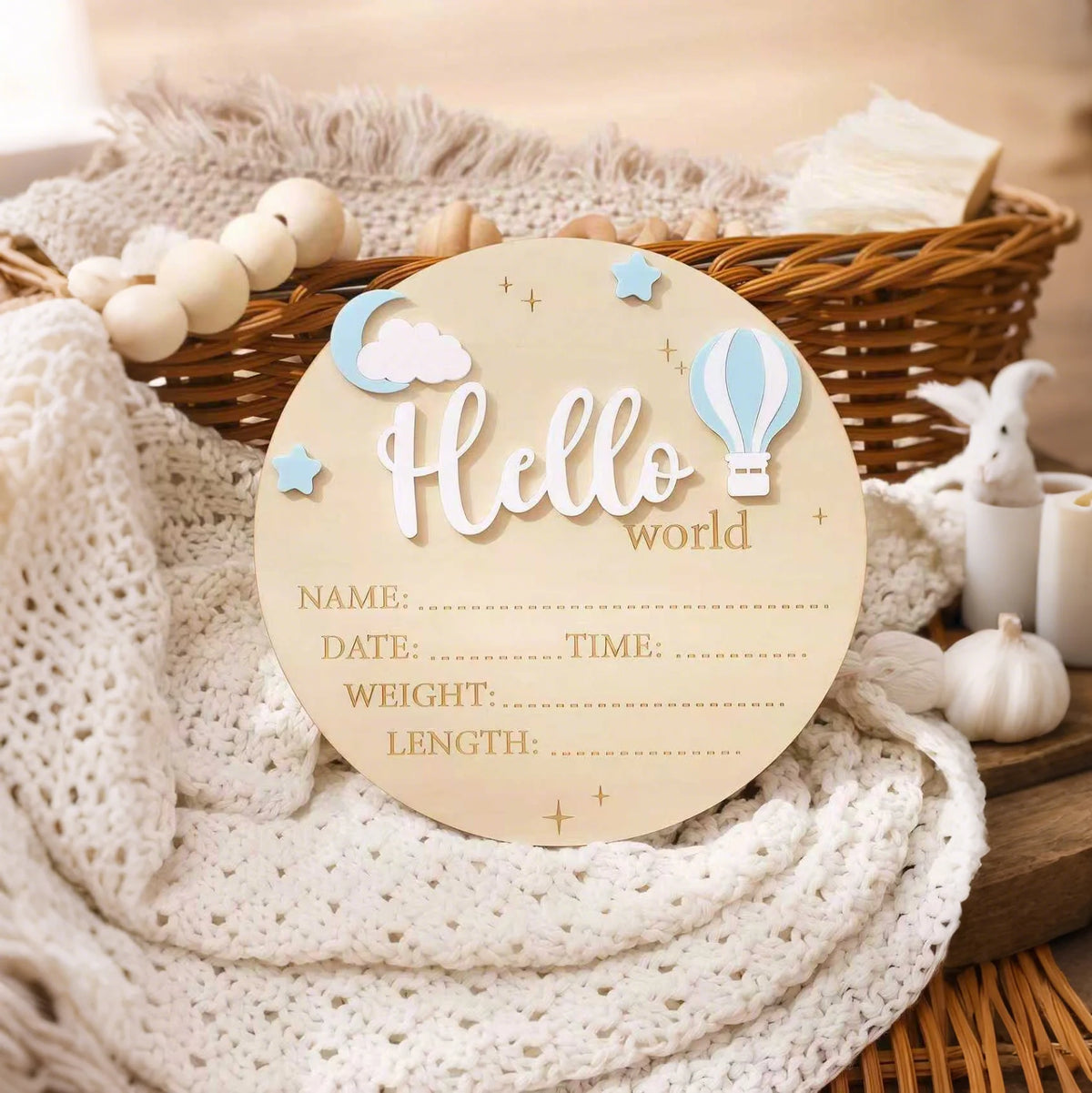 A round wooden birth announcement sign with the word Hello and a cloud and hot air balloon design, resting on a white knitted