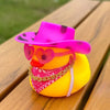A rubber duck shaped like a cowboy hat with pink and black patterns, wearing an orange heart necklace, sitting on a wooden su
