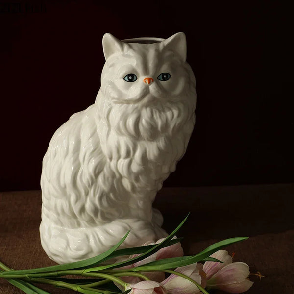 A white ceramic cat figurine with orange eyes sitting next to pink flowers.
