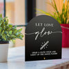 A black acrylic sign with white script that reads Let Love glow and features an illustration of a glow stick.