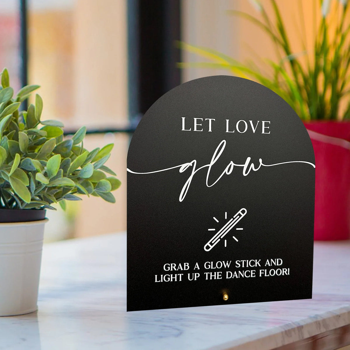 A black acrylic sign with white script that reads Let Love glow and features an illustration of a glow stick.