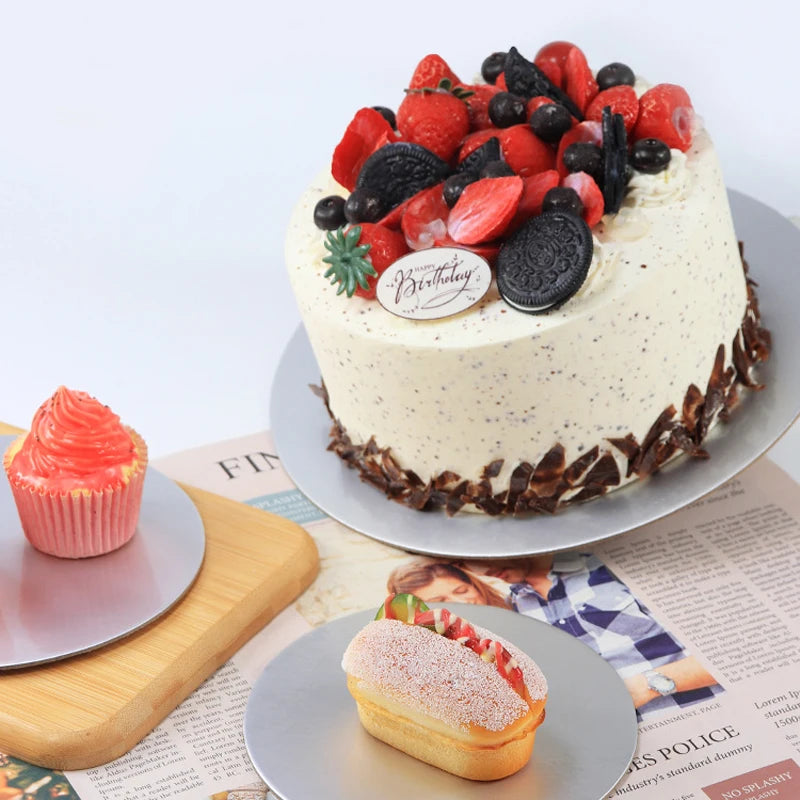 A white frosted cake decorated with fresh strawberries and blueberries is displayed on a silver cake board.