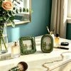 A vintage baroque resin picture frame in green holds a small glass bottle on a marble surface.