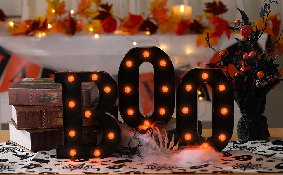 Un letrero de marquesina negro con forma de 'BOO' y luces naranjas brillantes sobre una mesa decorada con hojas de otoño.