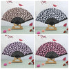 A leopard print folding hand fan with a black handle displayed on a wooden stand.
