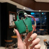 A hand holds a small green mini house handbag with a pink and red wrist strap.