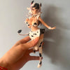 A hand holds up an adorable cow-girl miniature figure set featuring a white and black cow print outfit and pink hair.