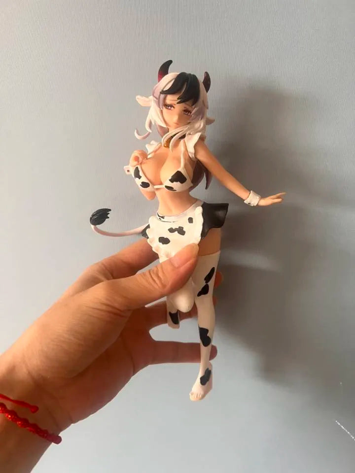 A hand holds up an adorable cow-girl miniature figure set featuring a white and black cow print outfit and pink hair.