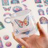 A hand holds a clear glass container filled with vintage floral window scrapbooking stickers.