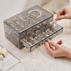 A clear acrylic jewelry organizer with three drawers is shown being handled on a white fluffy surface.