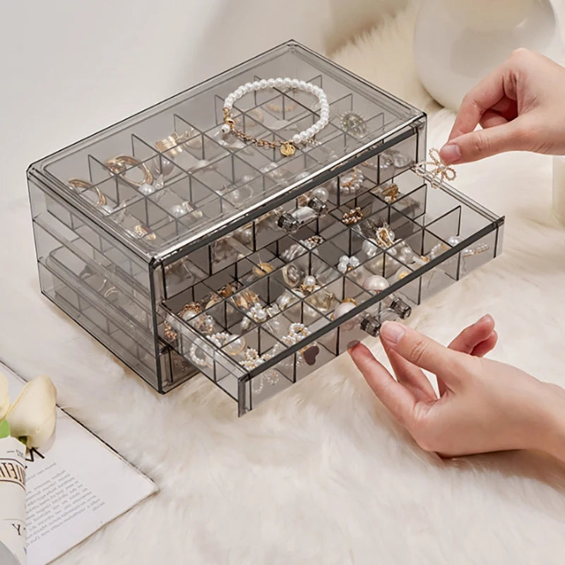 A clear acrylic jewelry organizer with three drawers is shown being handled on a white fluffy surface.
