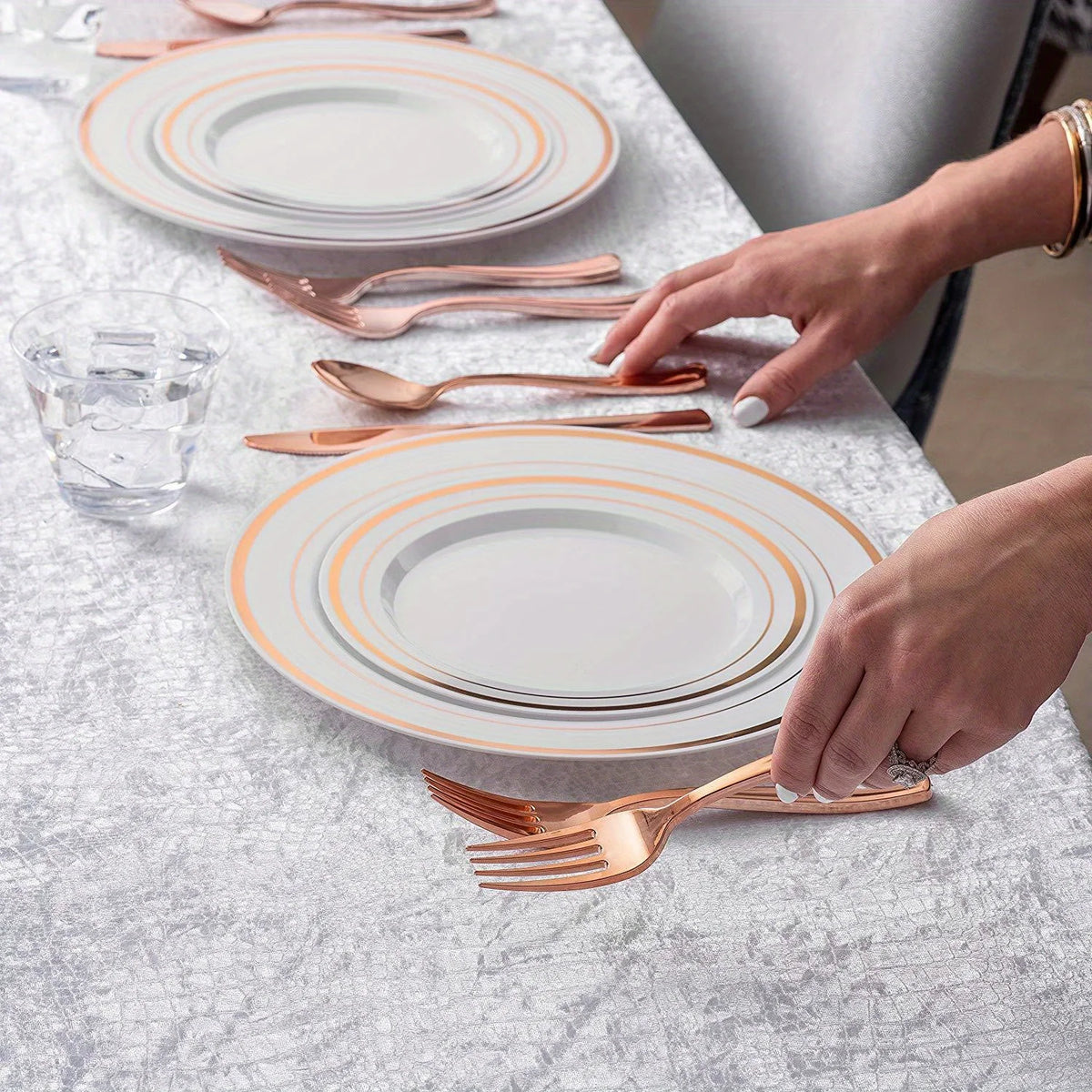 A hand places a gold disposable cutlery set onto a white plate on a silver-grey tablecloth.