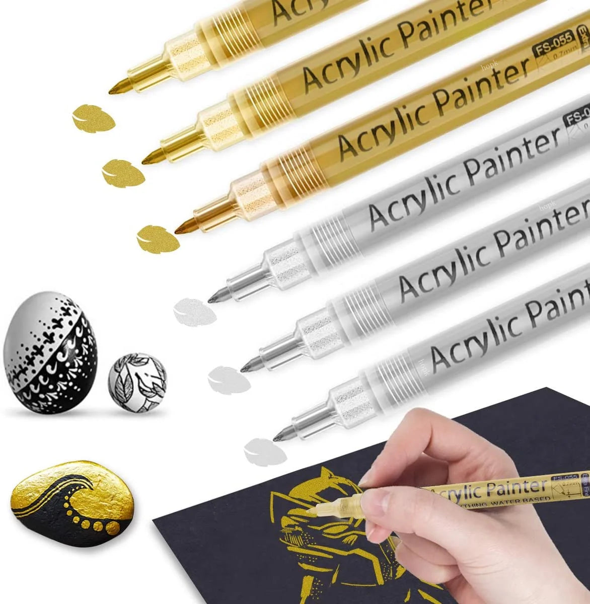 A collection of extra-fine point acrylic paint pens in various colors and finishes shown next to a hand painting on dark pape