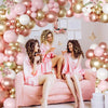 Four women in pink satin pajamas pose on a pink sofa surrounded by a rose gold and white balloon garland.