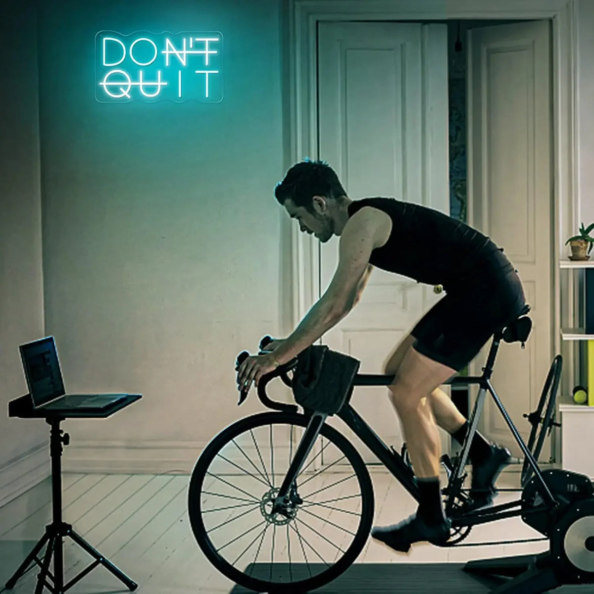 A man is riding a bicycle indoors in front of a neon sign that reads DON'T QUIT.