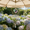 A gold heart-shaped pendant with a clear crystal hangs from a market umbrella amidst blooming flowers.