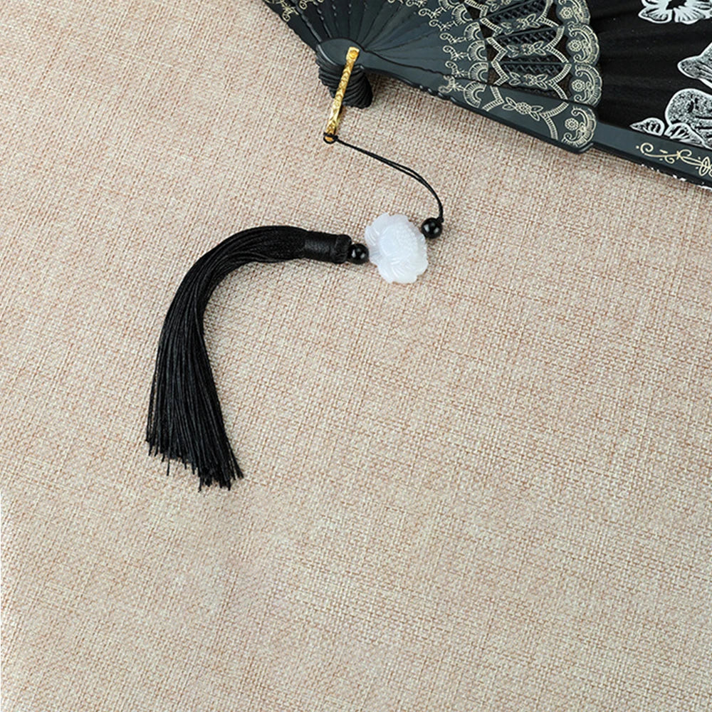 Black bamboo flower antique folding fan with a gold tassel and white pom-pom on a beige fabric background
