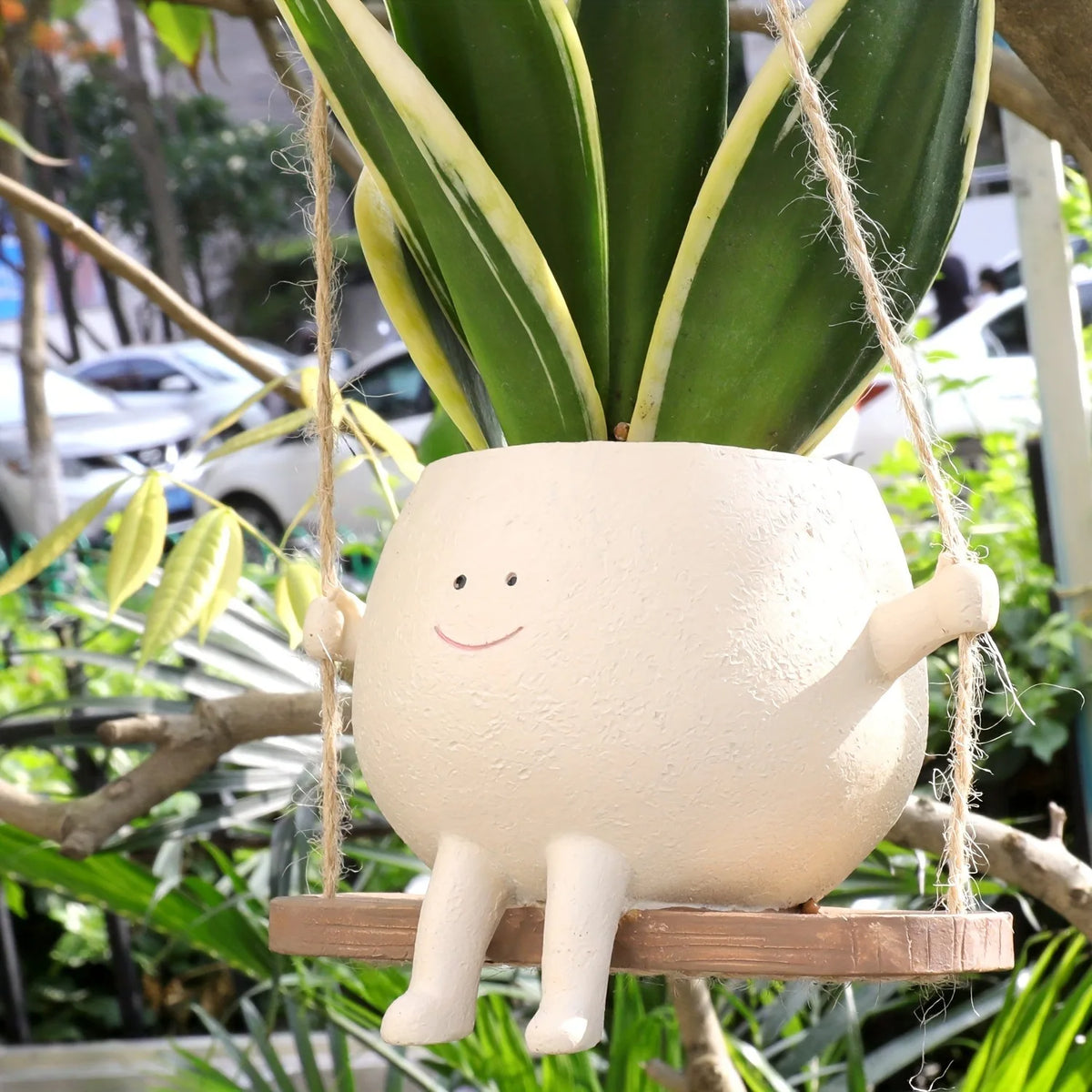 A white, smiling succulent planter hangs from a natural wood swing base amidst green foliage.