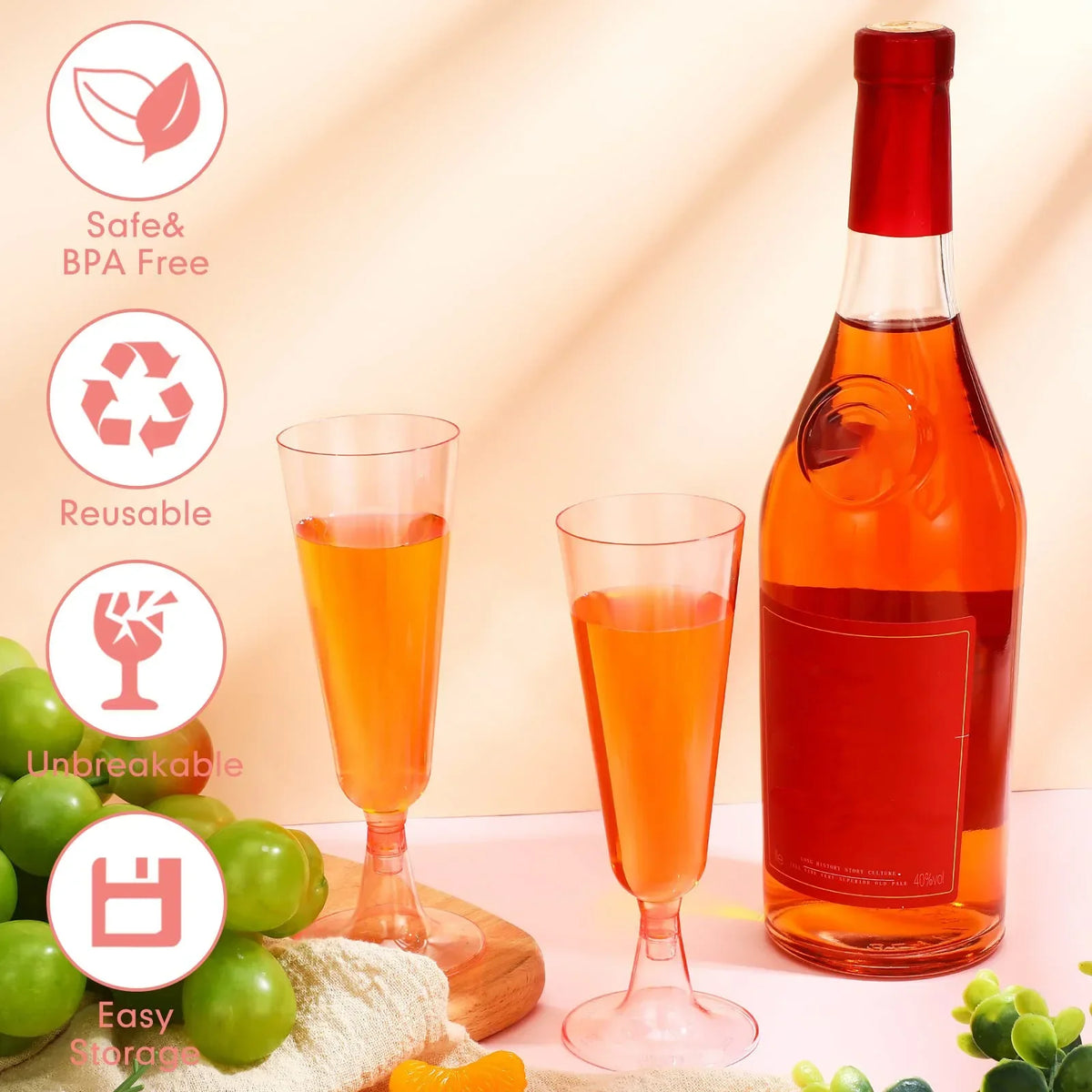 A set of two disposable champagne glasses with a gold finish are shown next to a bottle of orange-colored liquid and fresh gr