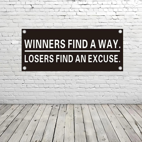 A black rectangular sign with white text that reads Winners Find a Way, Losers Find an Excuse mounted on a white brick wall.