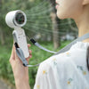 A person holds a white, handheld mini fan with a camera lens and a strap, aiming it toward foliage.