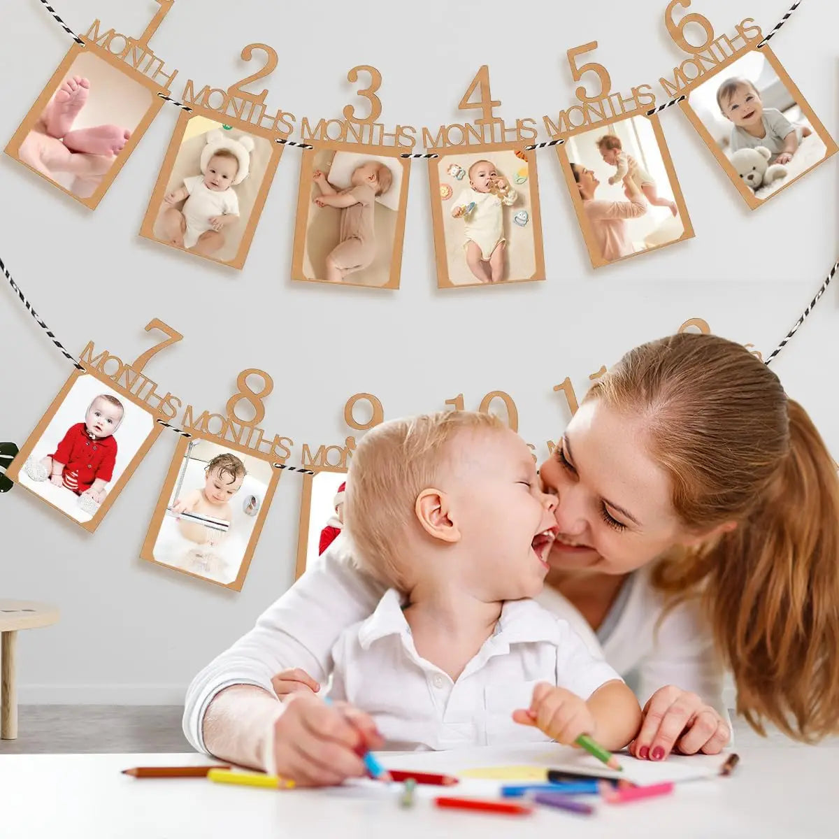 A woman is holding a baby while decorating a banner with monthly growth records for a baby's first birthday.