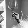 A hand holds a clear wine glass against a bright background, with a black and white art print visible in the foreground.