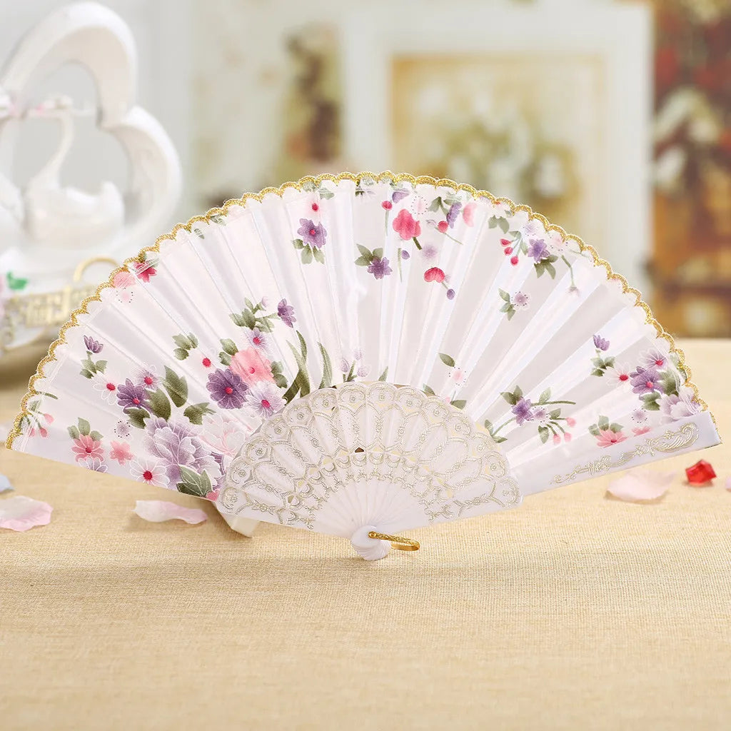 A delicate pink and purple floral lace hand fan with a gold edge resting on a beige surface.