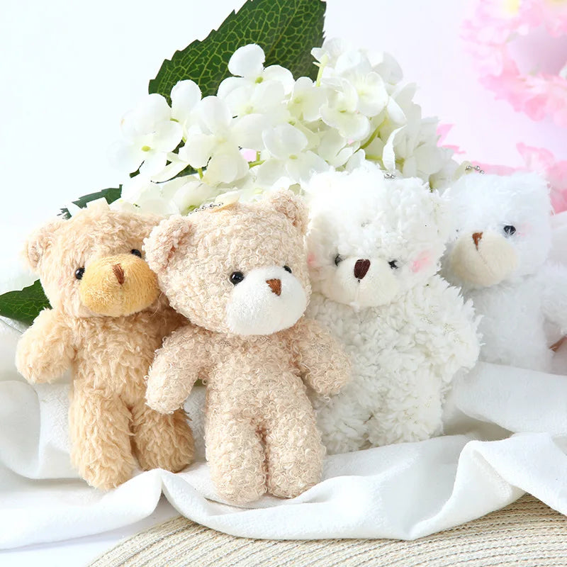 A set of three mini plush teddy bears in shades of tan and white with white flowers in the background.