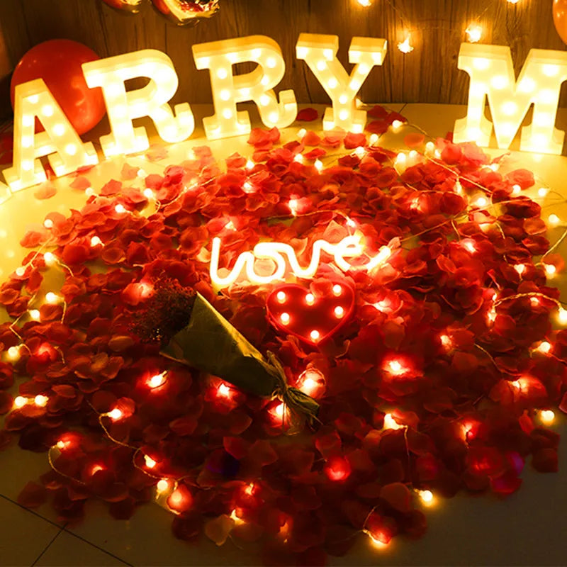 A close-up of red silk rose petals arranged in a circular display with warm string lights.
