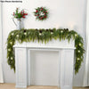 A green pine vine wreath is displayed on a white mantelpiece decorated with greenery and holiday accents.